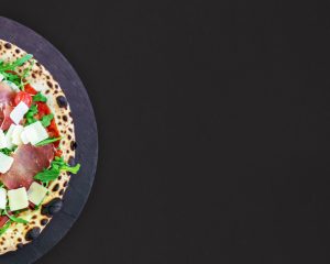 Une pizza alléchante, fraîchement sortie du four à bois, trône sur une assiette en ardoise. Sa pâte croustillante est garnie de jambon italien, de tomates cerises, de roquette et de parmesan râpé, offrant un contraste de couleurs et de textures appétissant. Le fond noir met en valeur la composition harmonieuse de la pizza et son aspect rustique authentique.