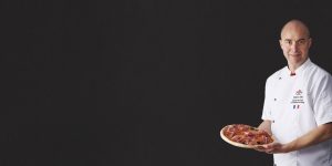 Grégory Edel, champion de France et vice-champion du monde de pizza, présente fièrement sa pizza au jambon italien. La pizza, garnie généreusement de jambon italien de qualité, de mozzarella fondante et de basilic frais, est cuite à la perfection dans un four à bois traditionnel.
